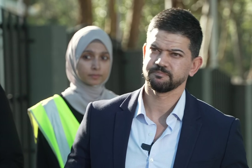 A man in a suit with a woman in a hijab behind him at a press conference.