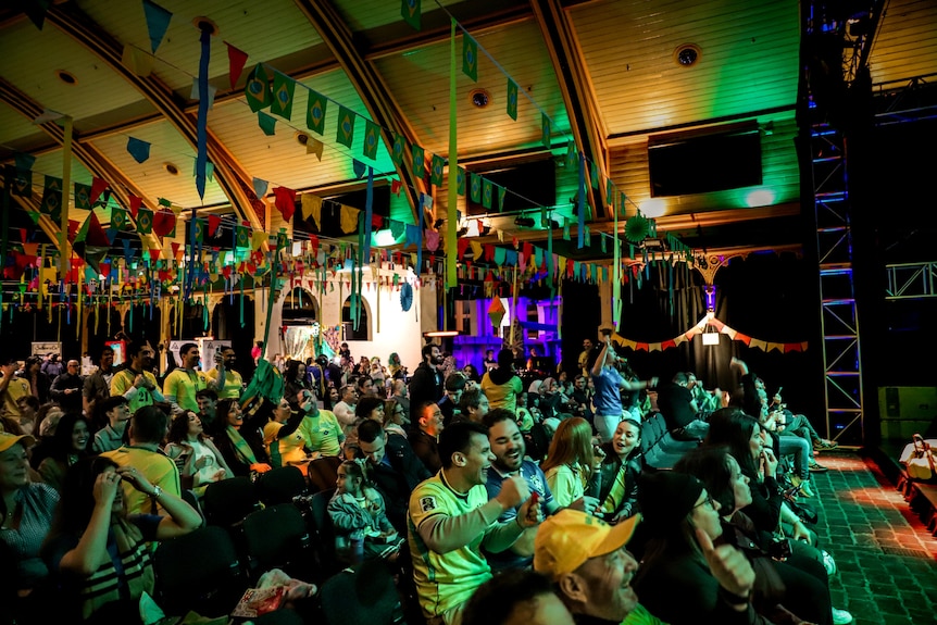 A large crowd of brazillian supporters wearing yellow and green celebrates in the glow of a projector screen inside event space 