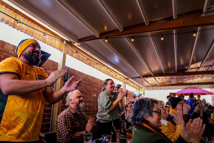 A beer garden of people cheering, some wearing yellow Australian outfits, amid wooden tables and brick wall