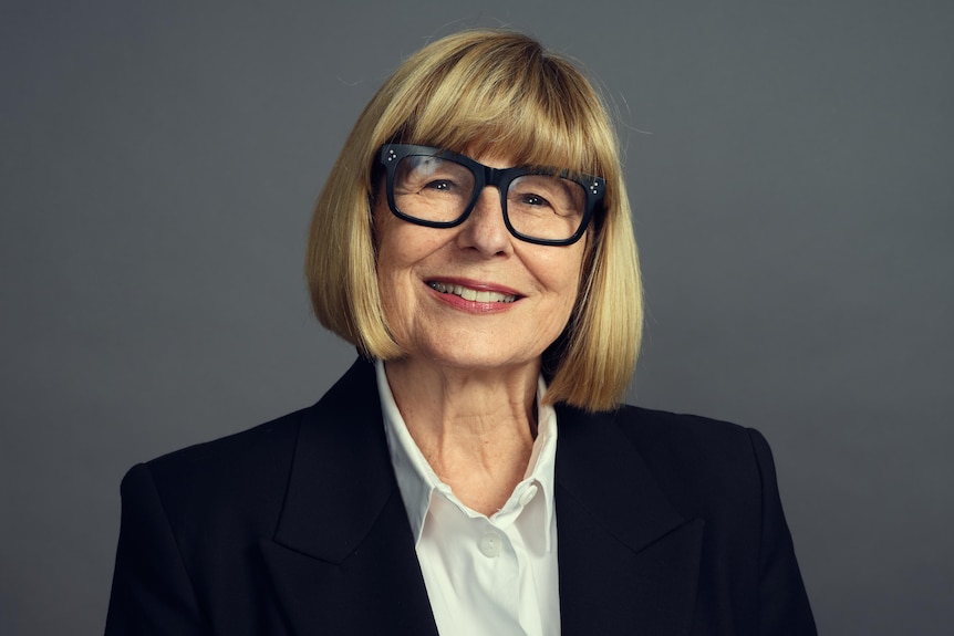 Woman in black suit jacket, short blonde hair, glasses