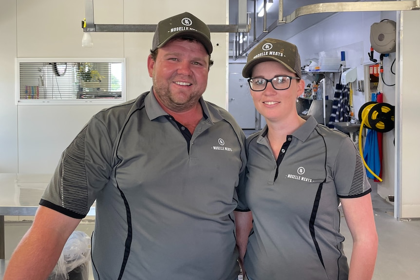 Man and woman with matching grey shirts and hats on stand next to each other in butcher shop smiling.