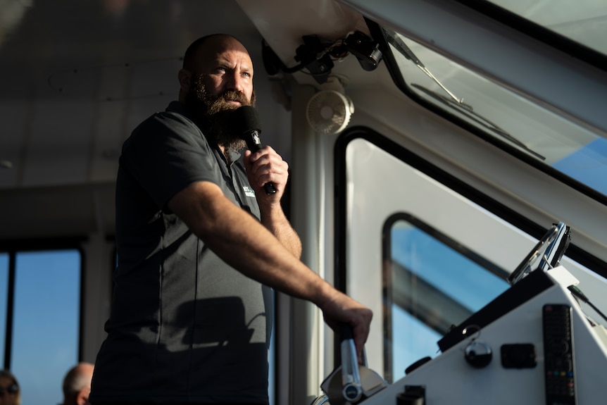 Spirit of the Coorong skipper and general manager Joel Hirsch steers a boat as he talks to passenger with a microphone