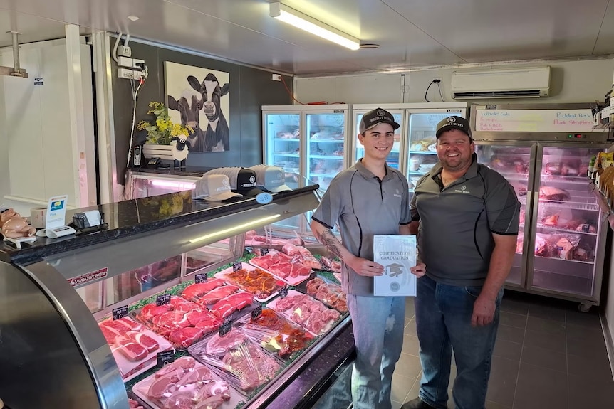 A young man with an older man standing in a butcher shop in front of the meat case, smiling looking into the camera.