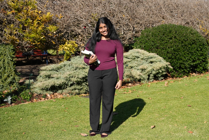 A woman wearing a maroon top and black pants smiles in a garden setting.
