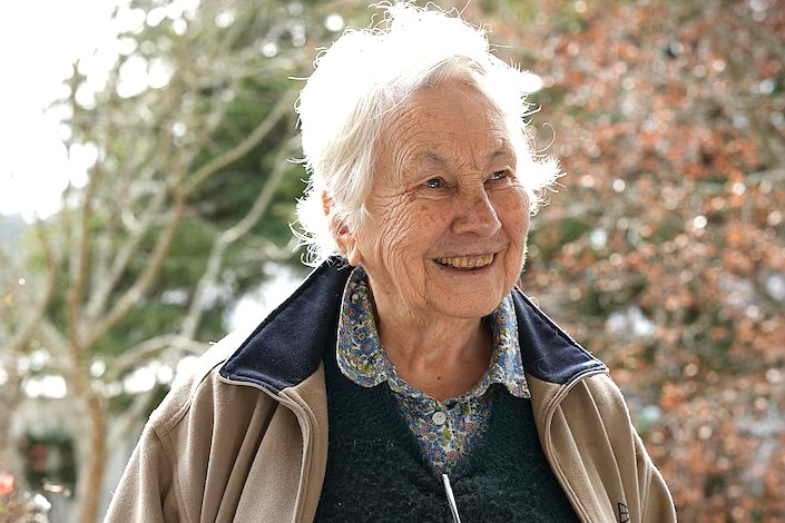 Elderly woman with white hair, smiling in front of garden.