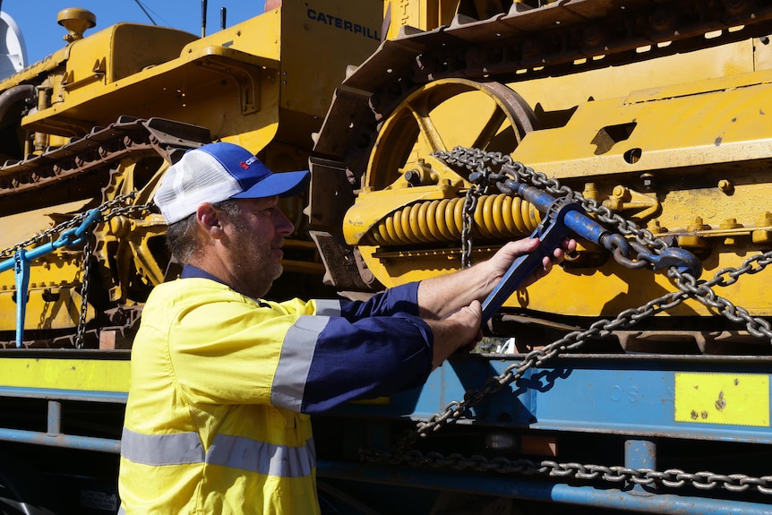 Truck driver adjusts chain holding down yellow bulldozer to truck bed