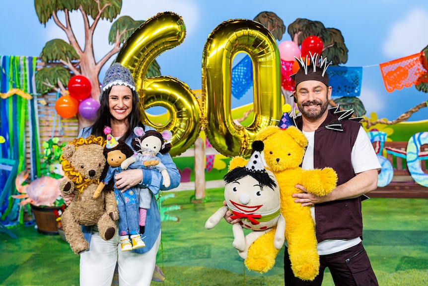 Two Play School presenters wear party hats and hold toys on colourful set. Behind them are gold '60' balloons