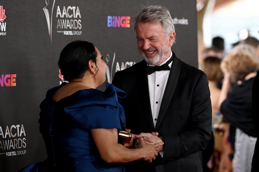 Sam Neill, wearing a tuxedo greets Deborah Mailman who is wearing a blue gown 