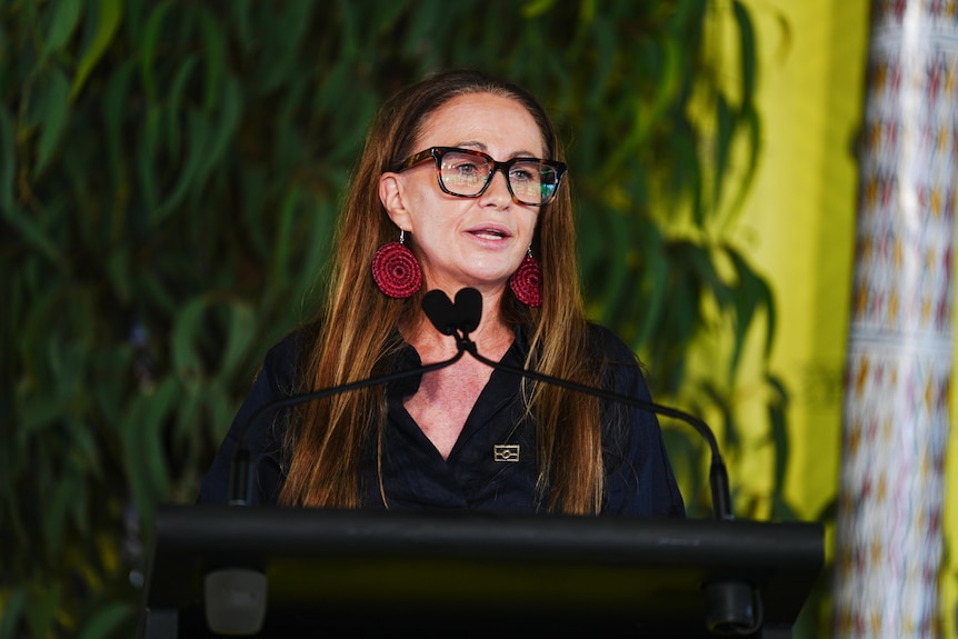 A woman with long hair and glasses speaks at a lectern.