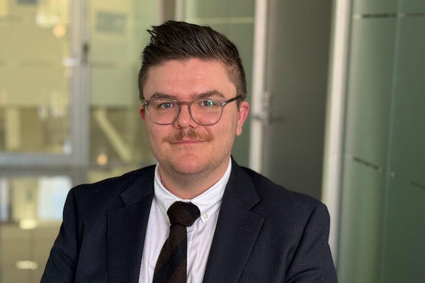 Kieren McKenzie, dressed in a dark tie and suit jacket, smiles as he stands in an office setting.