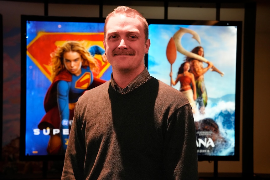 Mr Collins wearing a brown cardigan smiling as he stands in front of a movie poster of Moana next to Supergirl.