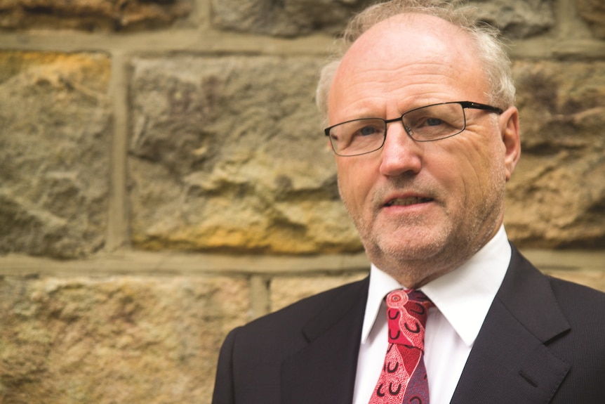 A man in a suit stands in front of a stone wall.