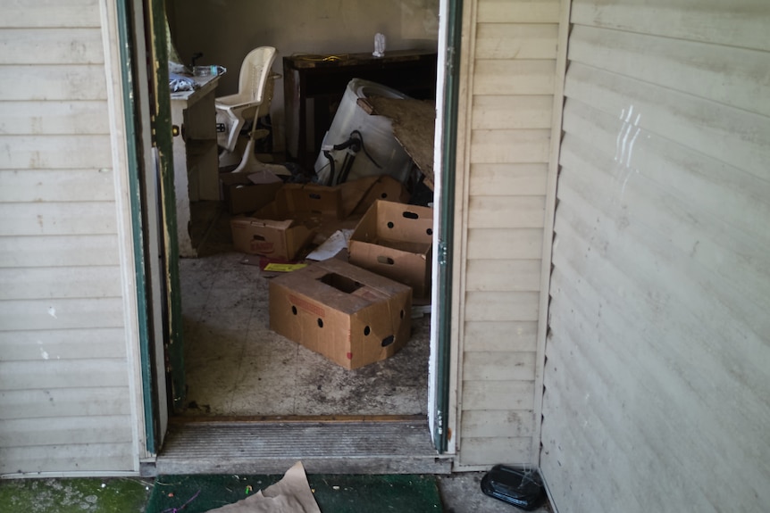 An open doorway shows the inside of a house with dirt on the floor and piles of cardboard boxes and miscellanous items