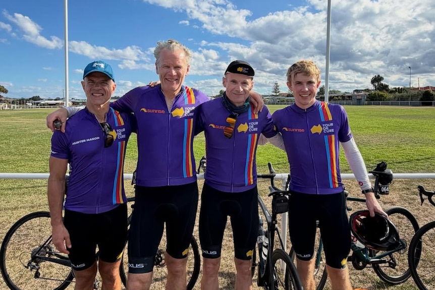 Four men stand arm in arm wearing matching purple cycling gear, their bikes behind them. They stand at football field, blue sky