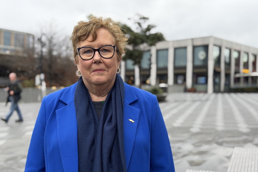 A woman with short hair and glasses wears a blue scarf and blue blazer.