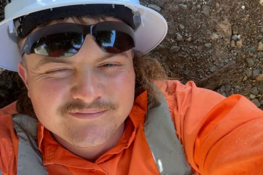 Man with blonde hair, wearing high-vis, sunglasses, hard hat, living on ground winking.
