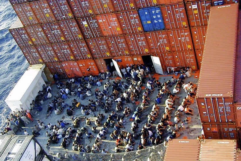 An aerial view of people sitting in rows. Shipping containers are stacked next to them.