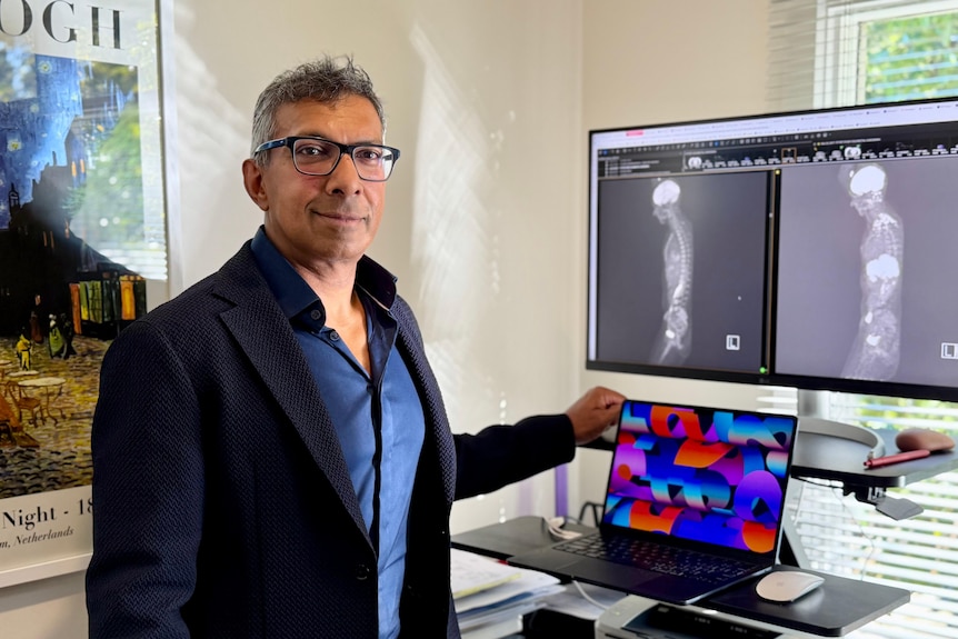 A man in a suit stands in front of a cumputer and some scans of the human body.