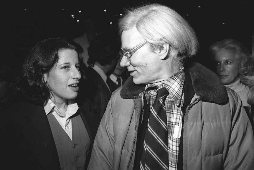 Black and white photo of a young Fran Lebowitz in conversation with Andy Warhol at a party in New York in 1977.