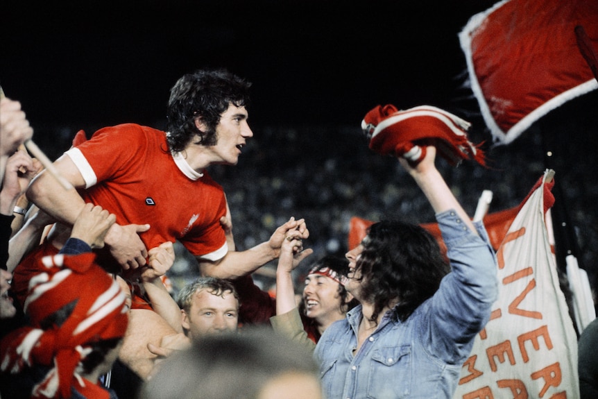 Celebrating fans carry a dark-haired player in a red shirt and with a 70s hairstyle and sideburns.