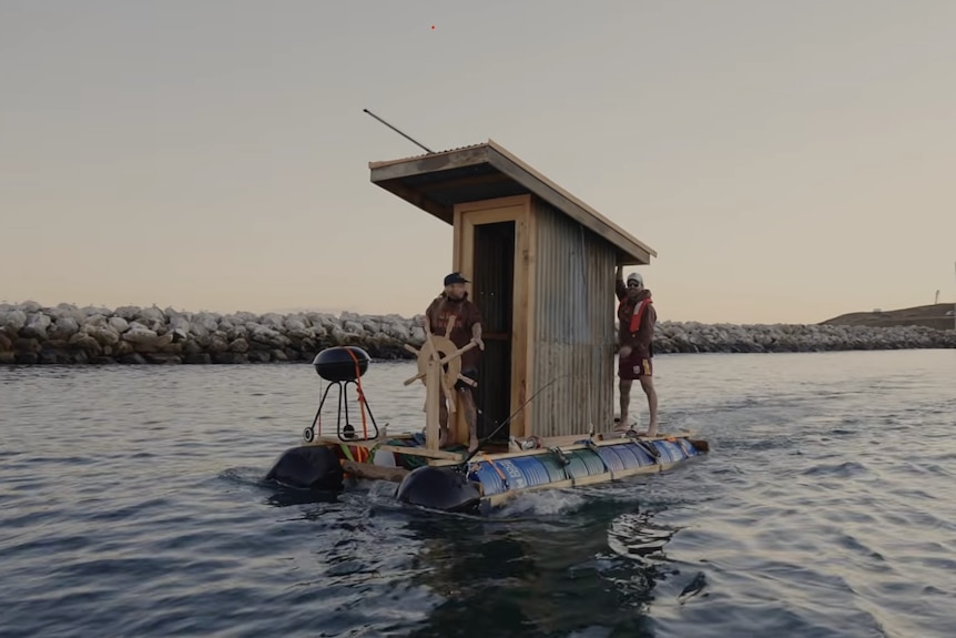 Two men aboard a barge with an old fashioned toilet and a barbecue on it
