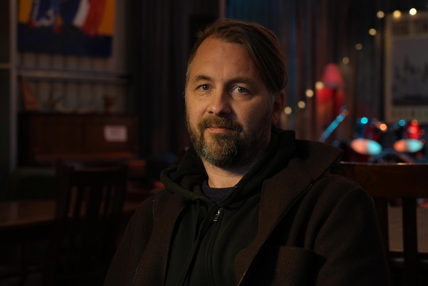 A man sits in dim light looking at the camera, in front of a bar with coloured lighting
