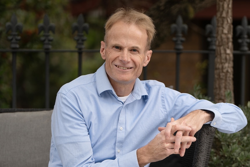Professor Scolyer sits leaning on his elbow in an outdoor seat. He wears a blue-collared shirt and jeans and is smiling