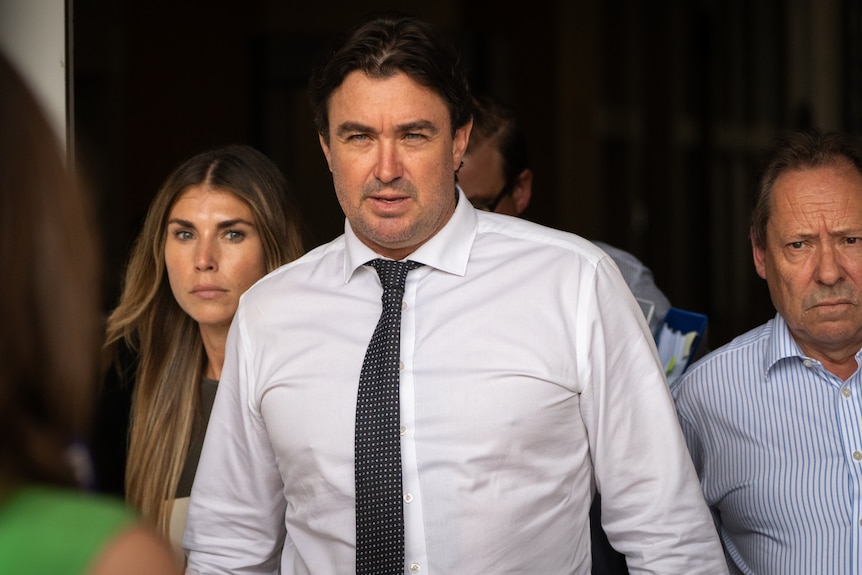 White man in a white button up shirt, gray tie walking out of building, with woman to his side and older man to his other side