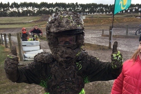 A young boy in a motorbike helmet is giving a thumbs up while covered in mud.