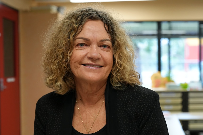 A woman wearing a black blazer sits in a classroom with windows and desks behind her. She has curly light brown hair, is smiling