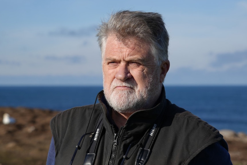 A man with grey hair and a white beard stands in front of the ocean, while looking away.