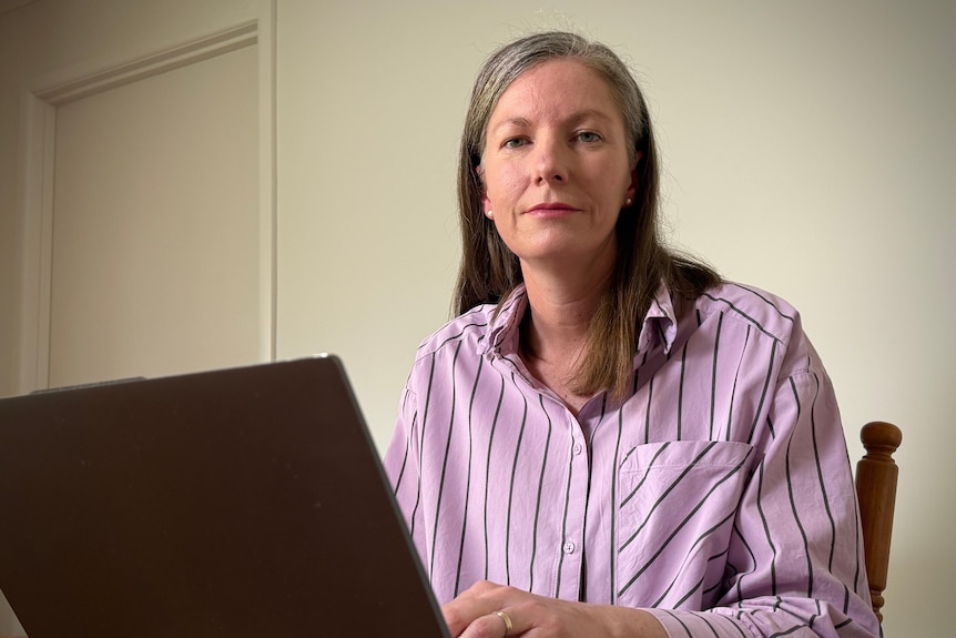 A woman has grey hair and wears a pink striped top, she sits at a laptop looking at the camera