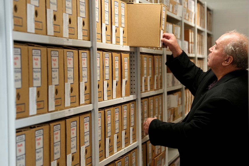 A man wearing a black suit jacket reaches for a file in an archive room.