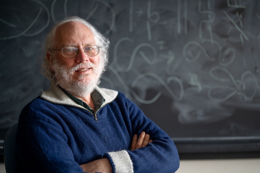 Bearded man with white hair and glasses, smiling with his arms crossed in front of a whiteboard with maths equations in chalk.