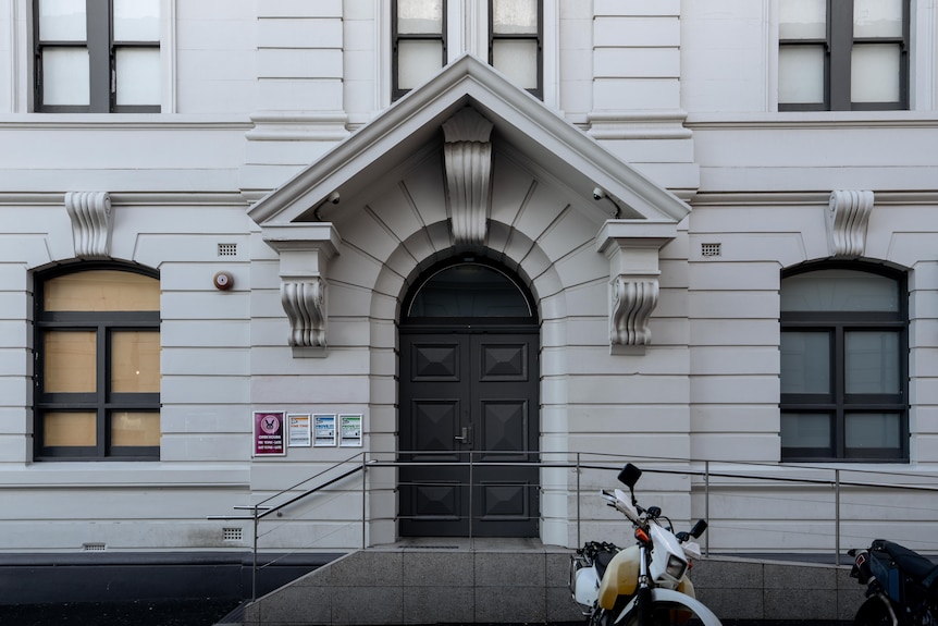 A light grey, almost white, heritage building with motorcycles out the front and signage by the door near grey-rimmed windows