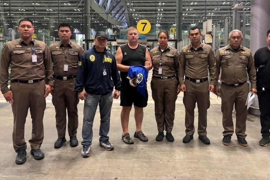 Eight police officers stand in a line next to Simon Carman at an airport.