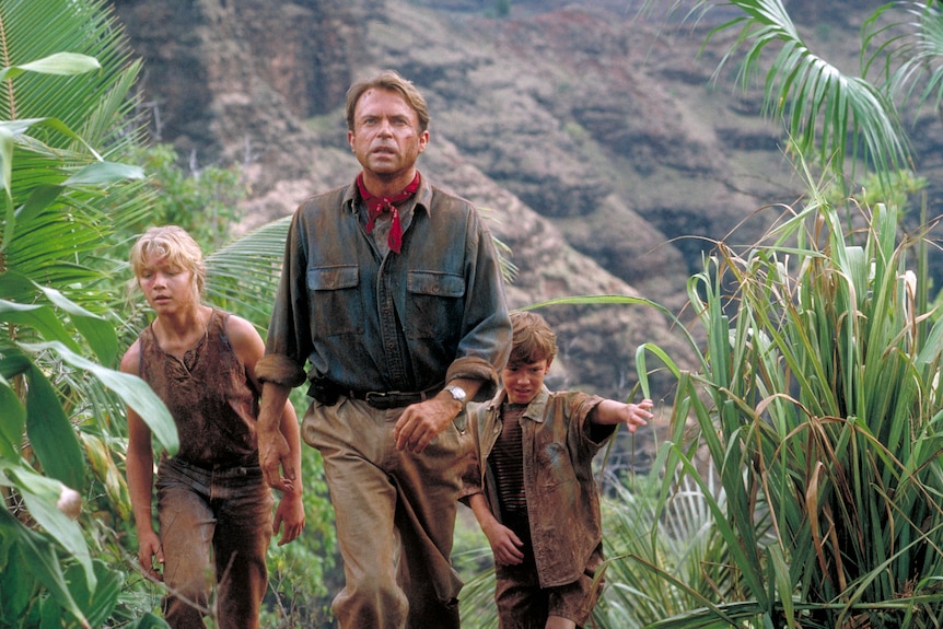 Still from the movie Jurassic Park Sam Neill walks through jungle with two child actors 