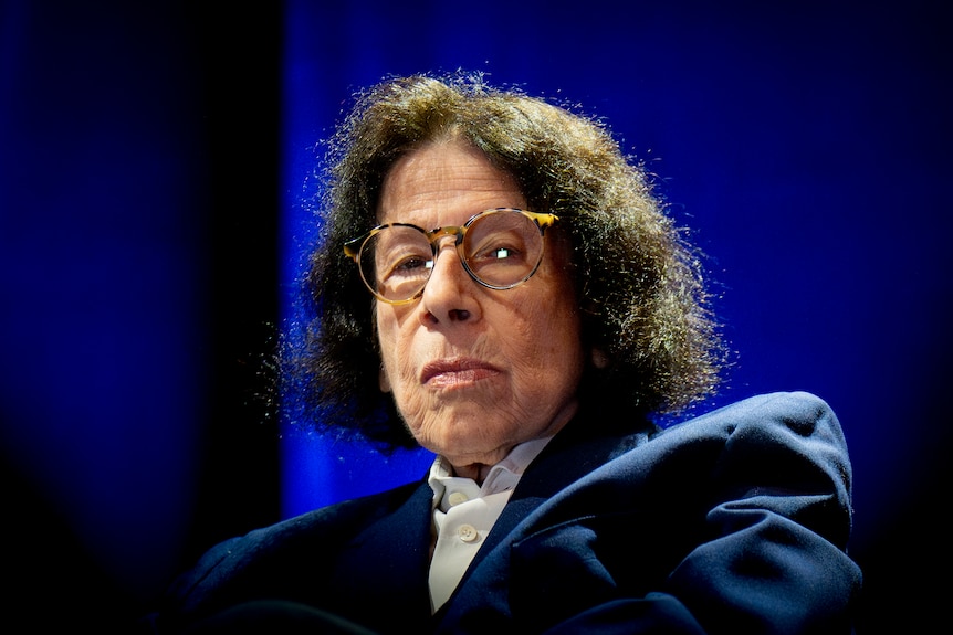 Close up shot of Fran Lebowitz staring down the lens, on stage against a blue curtain wearing a navy jacket and white shirt
