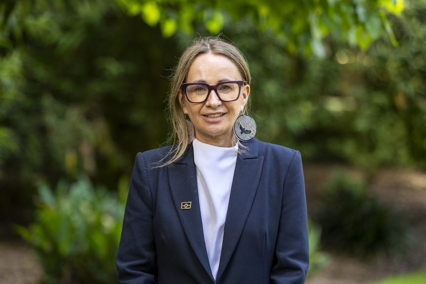 A woman with light brown hair wearing glasses, a white shirt and a blue blazer smiling into the camera.