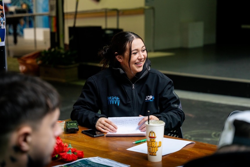 A young woman wearing a black jacket sits at a table with pen and paper, smiling to someone out of frame.