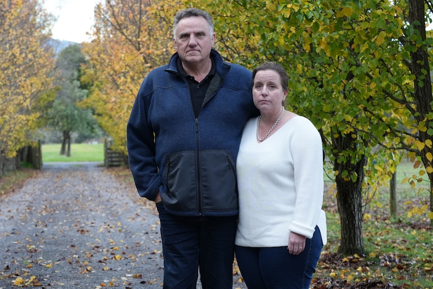 A man and a woman stand in a driveway lined with deciduous trees.