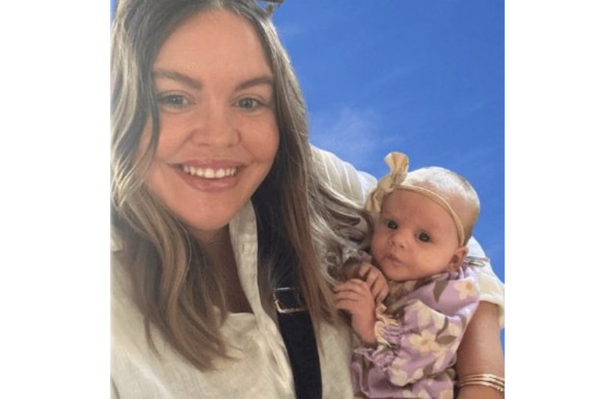 A woman with brown hair and sunglasses on her head smiling holding a small baby 