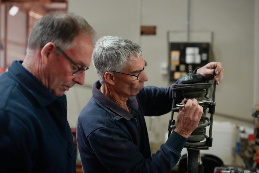 Two middle aged men working on car parts on bench in mechanic workshop