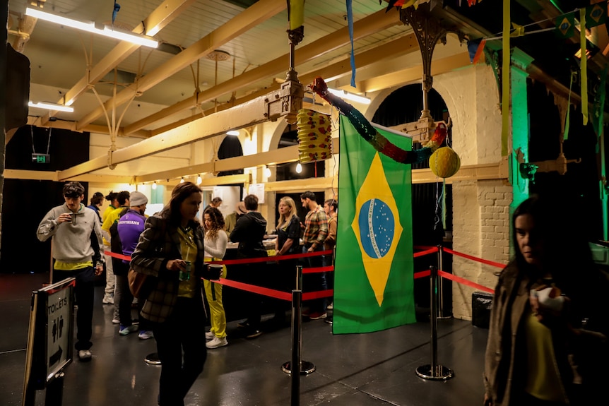 People walk from an indoor bar past a large Brazilian flag inside a dimly lit room