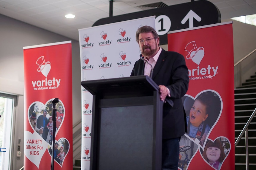 A man stands at a podium with red signs behind him with a red banner that says "Variety".