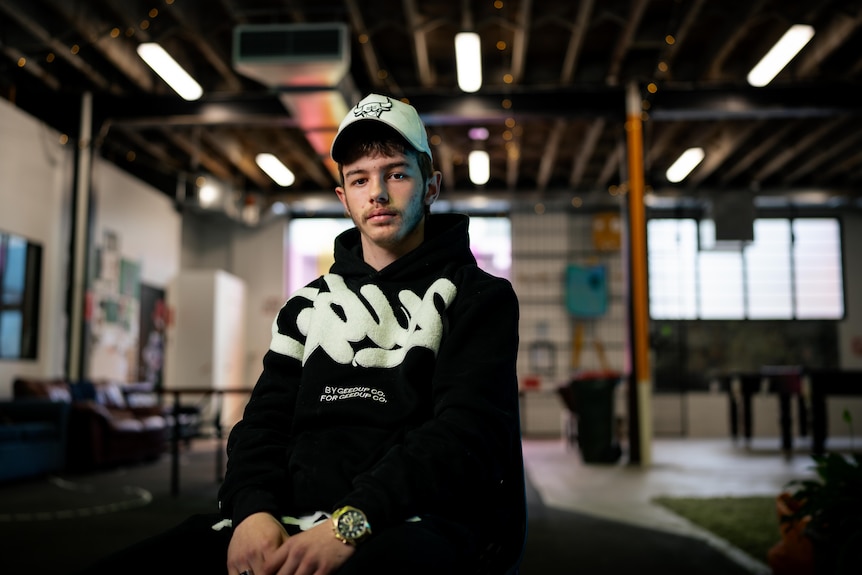Latrell, wearing a black hoodie with white writing and a white hat, sits on a chair with a serious expression.