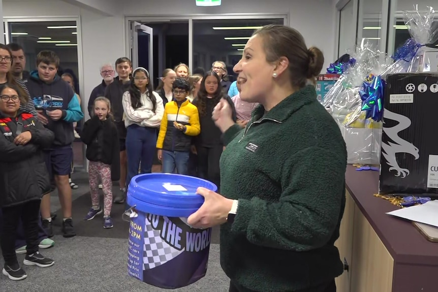 A woman with a green jumper holding a blue bucket while surrounded by a large group of people of mixed ages and genders.