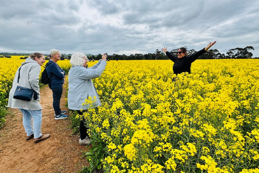 A woman poses enthusiastically in a canola field with three others watching on