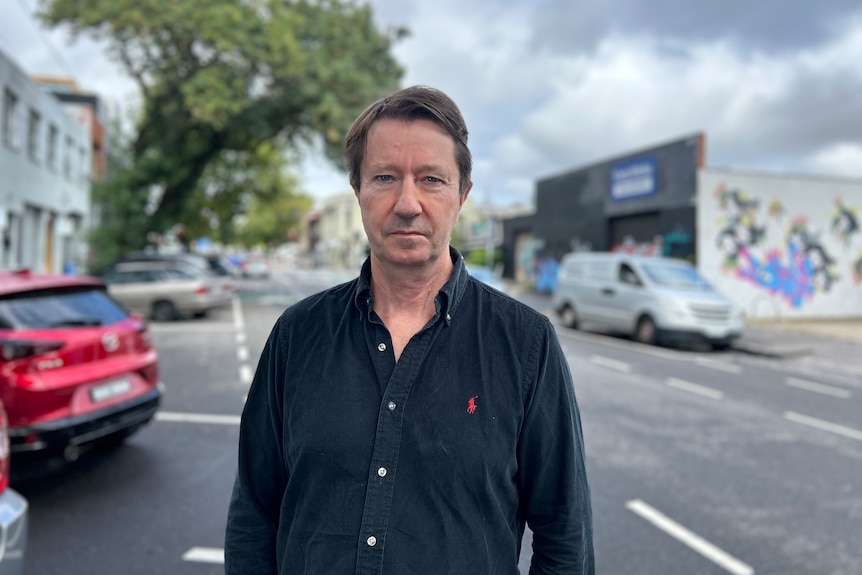 A man wih brown hair in a black collared shirt stands in a street near parked cars on a cloudy day.