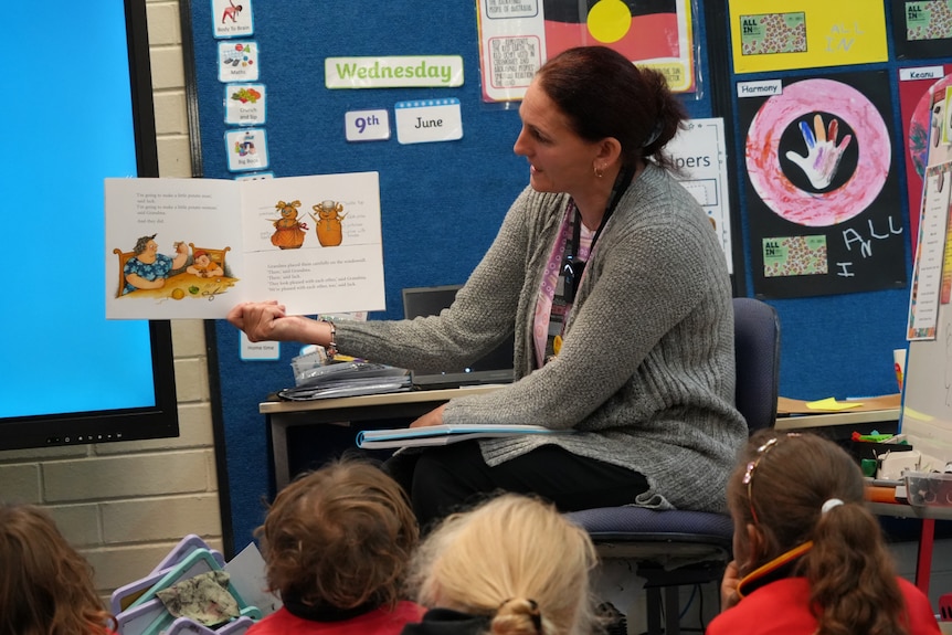 Children seen from behind sitting on the ground in a classroom. An adult a the front is facing the children and holding a book.